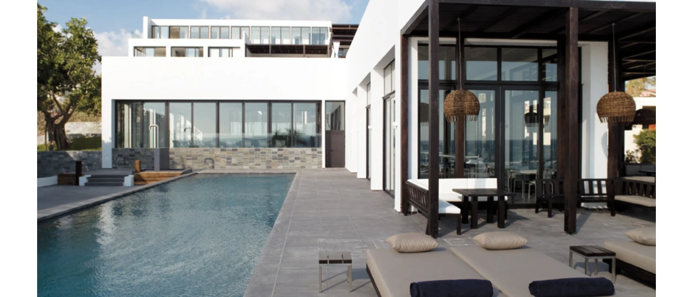 Rectangular swimming pool next to a terrace lined up with cushioned loungers, next to a modern building with full-length windows