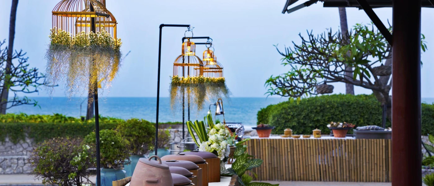 Outdoor table stacked with pots with closed lids, under rustic lantern lights with the ocean in the background