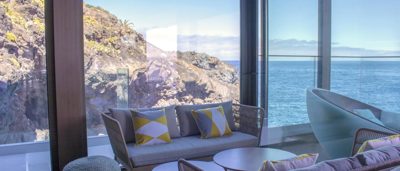 Cushioned seating area and coffee table next to full-length windows overlooking the sea and cliffs