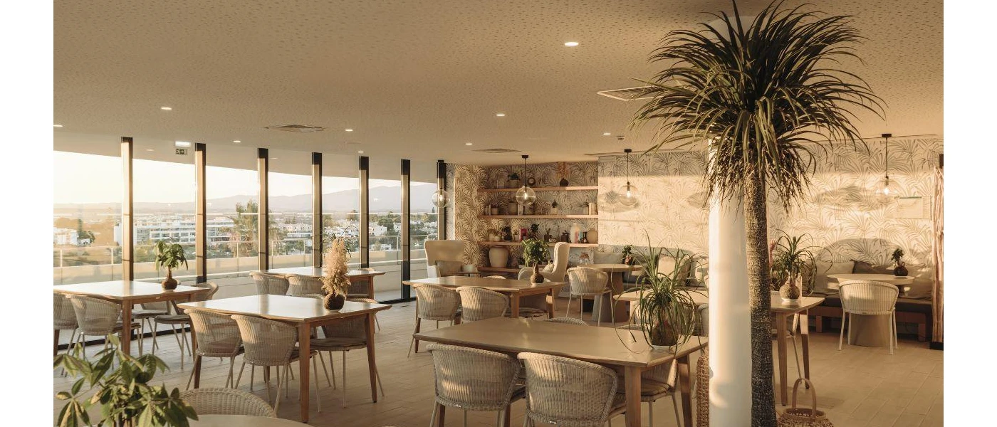 Restaurant with wooden floors, chairs and tables, a cream papered wall with a palm design, and potted palms and shrubs, plus floor-to-ceiling windows