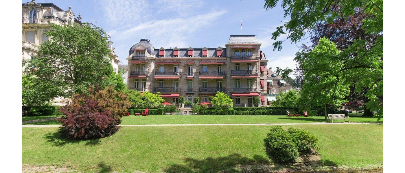 Grand, multi-storey buildings with iron balcony railings and red shutters surrounded by lawns and trees under a blue sky