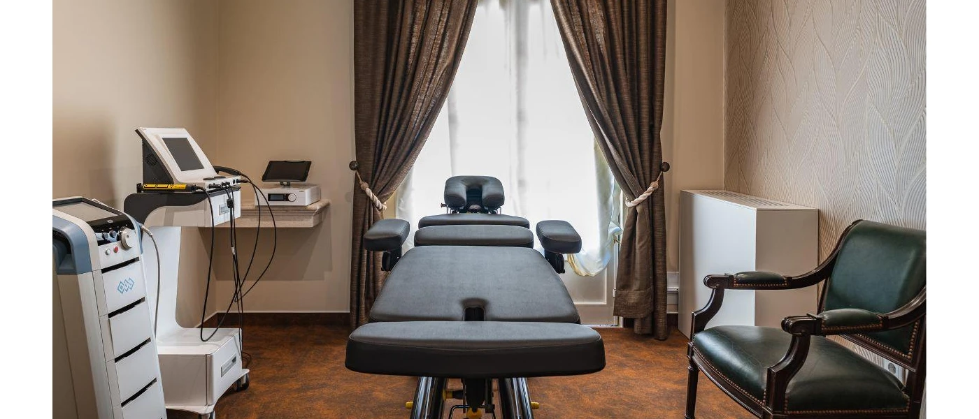 Neutral room with a wooden floor and dark drapes, with a black leather medical chair, machines and wooden chair