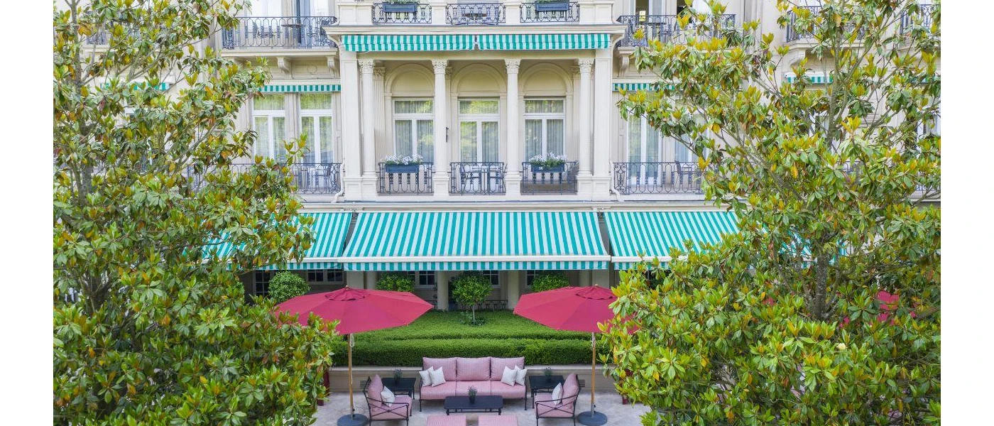 Grand facade on a mansion-style building, with a green and white-striped awning, red umbrellas and trees to the front