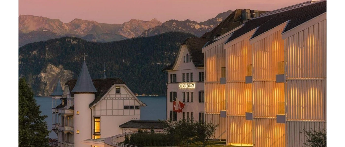 Chenot Palace at night, all white buildings and towers with grey turrets, with Swiss flags on the entrance and Lake Lucerne and the Alps in the background