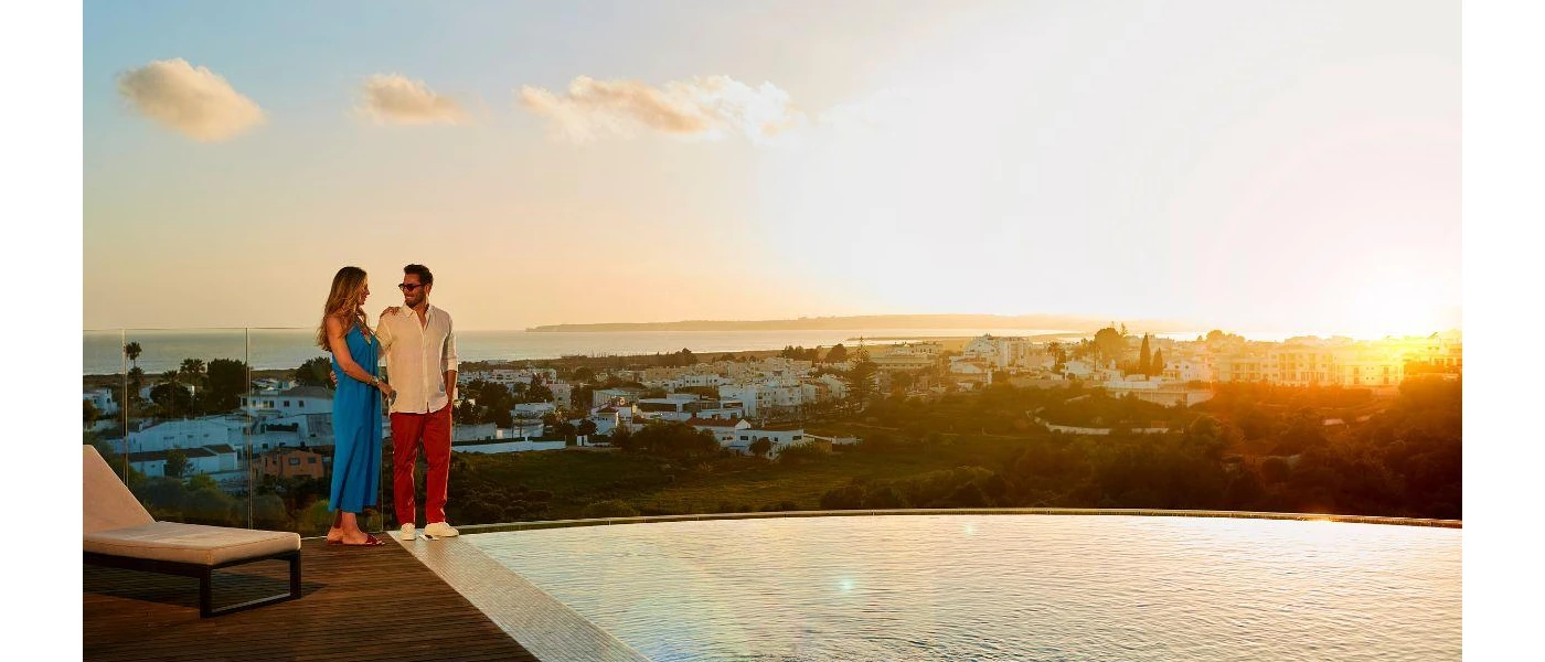 Woman in a blue dress and man in red trousers and a white shirt smile at each other on a glass-fronted rooftop as the sun sets in the background