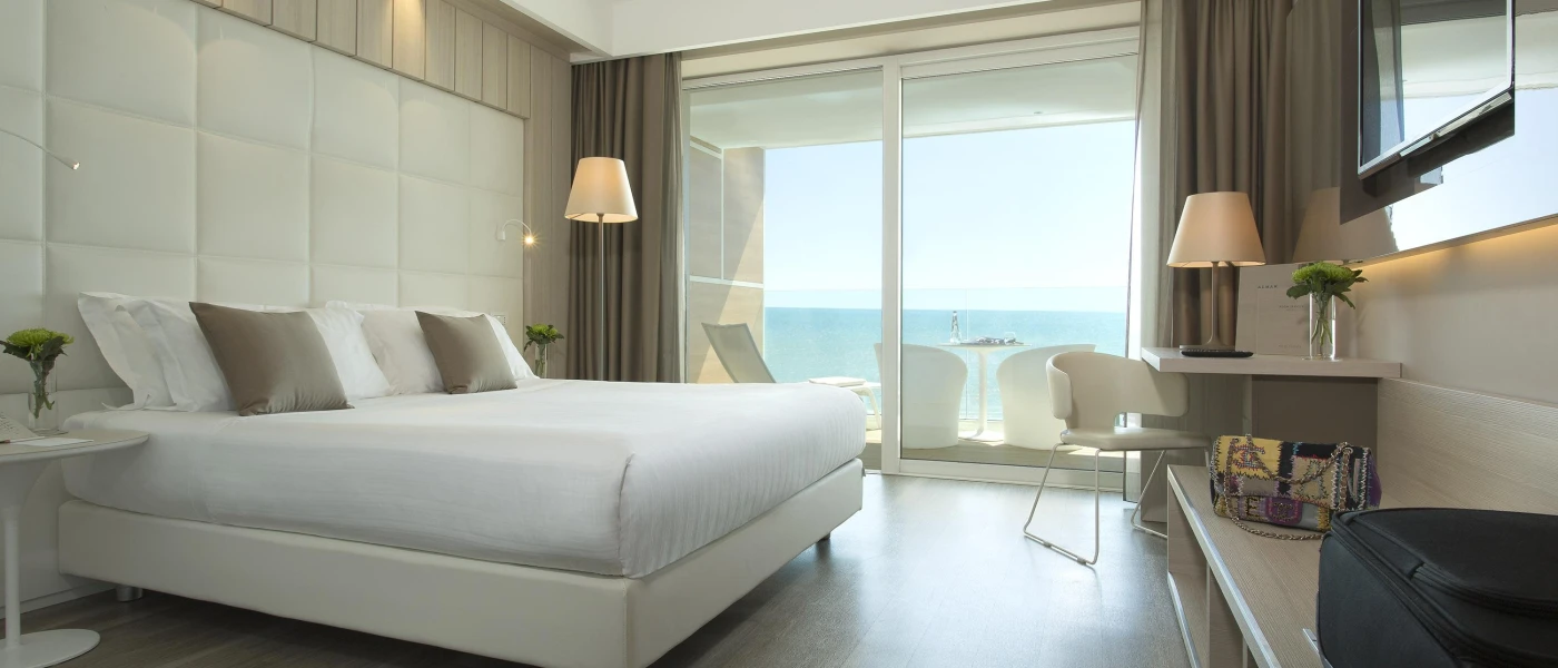 Hotel room with white bedding, wooden flooring, a neutral padded headboard and neutral lighting and cushions, with floor-to-ceiling windows overlooking a balcony
