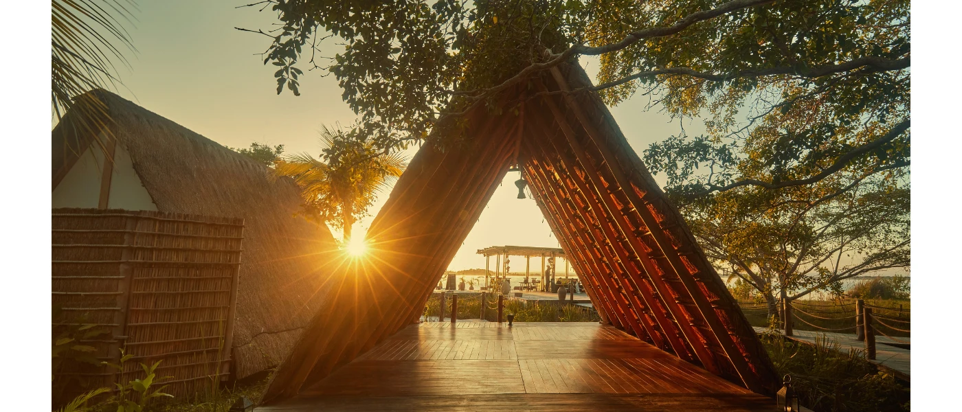 A-frame thatched structure opening onto a wooden deck surrounded by greenery as the sun sets in the background