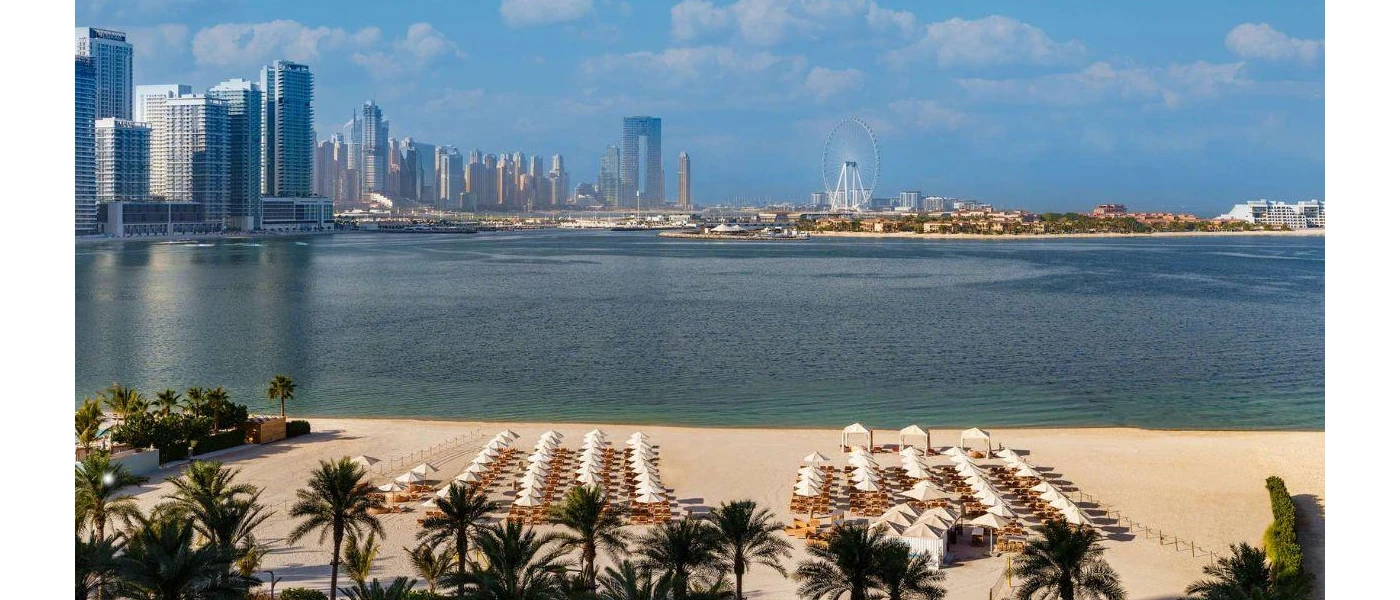White-sand beach neatly lined with white parasols and loungers, backed by palm trees and with a view of the sea and Dubai skyline
