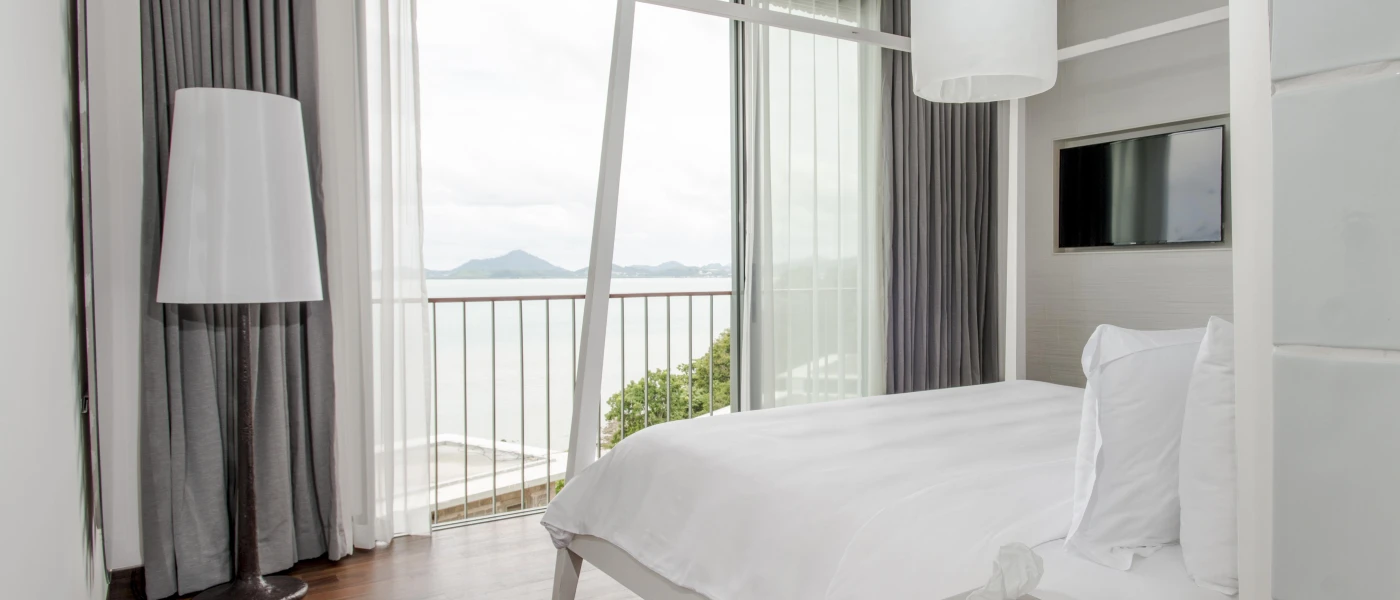 Hotel room with white bedding, grey curtains, white lamp shades and a full-length window with sea views