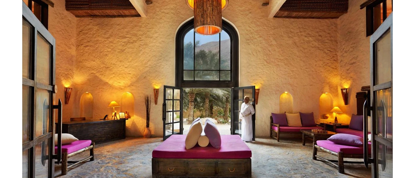 A lobby area with buttermilk stone walls, lantern-like lighting, dark-wood chairs with red cushions, and a staff member in traditional dress standing at the entrance
