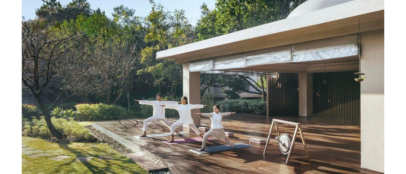 Three people in loose fitting white shirts practice yoga on a wooden deck, overlooking a sunny garden