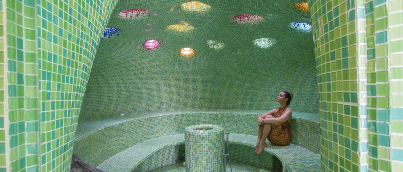 Woman relaxes in a steam room with intricate mosaic tiled design, including cut out ceiling sections in different colours
