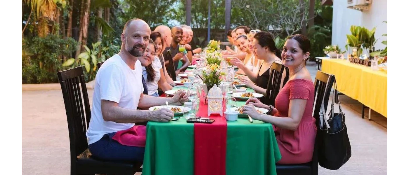 Smiling group pose for a photo while sitting around a communal dining table with a green and red tablecloth and tropical greenery in the background