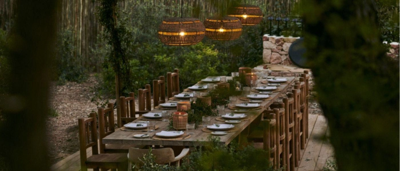 Outdoor rustic wooden table and chairs surrounded by trees and lit by lanterns above