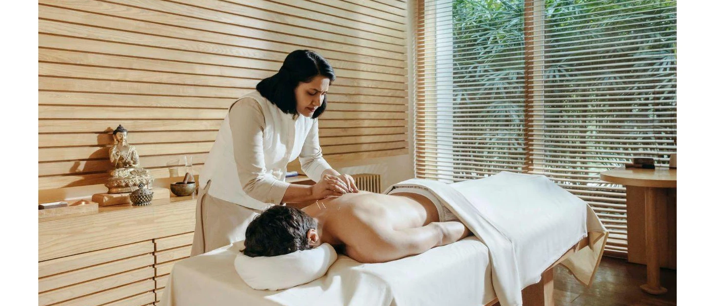 Man lies on his back draped in a white towel as a therapist in white massages him, the treatment room is wood-panelled and has a Buddha statue and sound bowl on a table 
