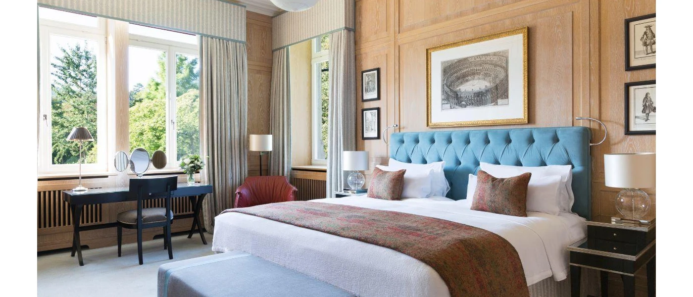 Elegant hotel room with teal headboard, white bedding, a dark-wood dressing table, and pictures of historical images on the wall