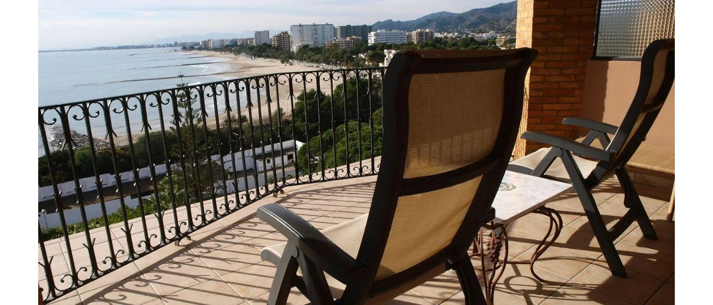 Black wrought-iron balcony with two chairs and a small table overlooking the sea and a golden beach