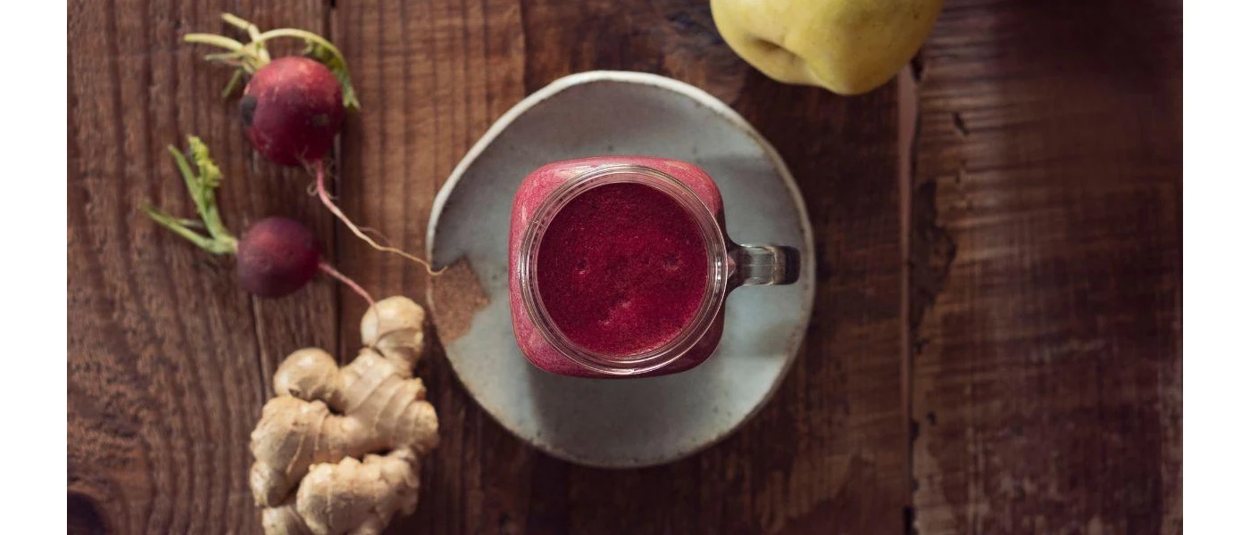 Jar of bright pink liquid surrounded by radishes, ginger and an apple