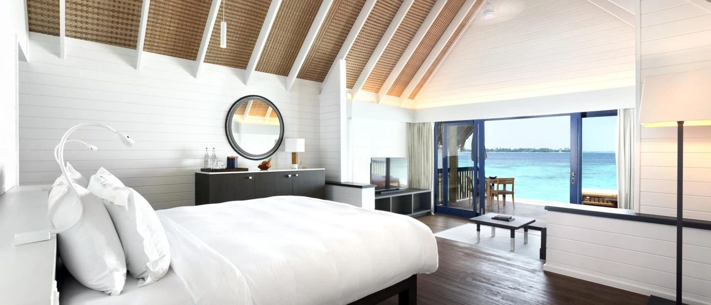 Minimalist villa bedroom with white bedding, a wood-beamed ceiling, dark-wood floors and a dressing table, with open doors showing off the ocean views