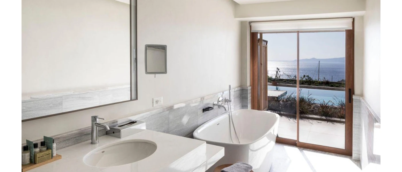 All-white bathroom with a bathtub and large wall mirror, and floor-to-ceiling windows overlooking shrubs, a pool and the sea