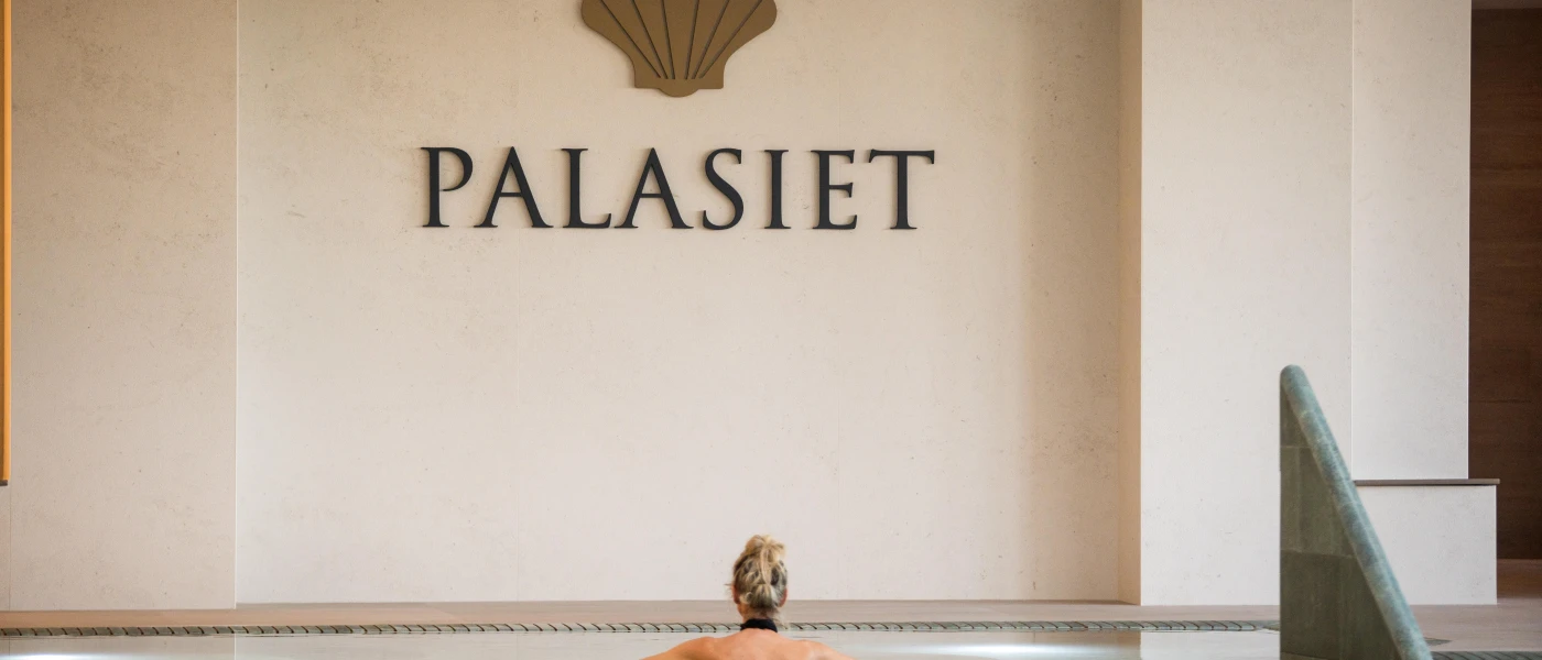 Woman swims in an indoor pool under a cream wall reading 'PALASIET'