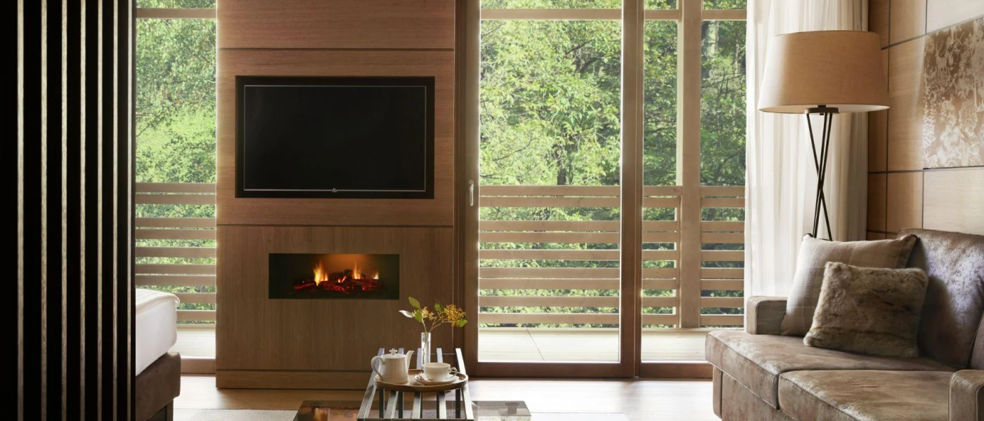 Living area with a plush brown sofa, glass coffee table, wood-panelled wall with a TV and fireplace and full-length glass doors overlooking a forest