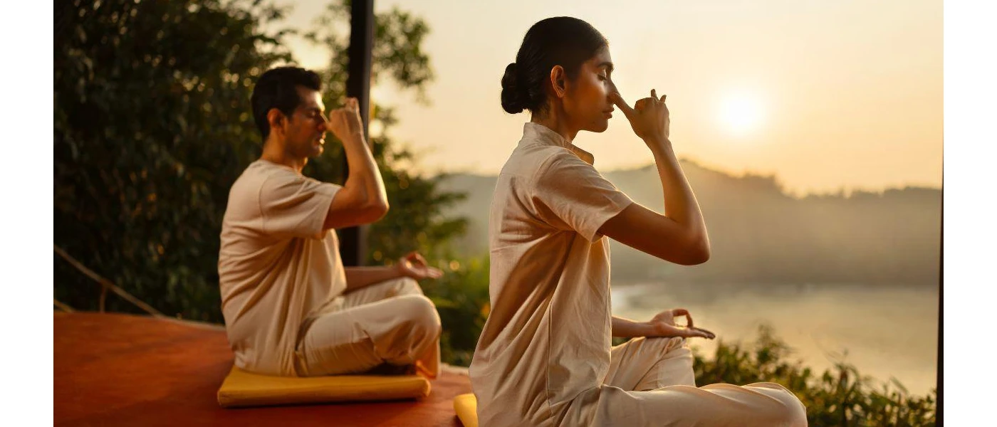 Two people in light, loose clothes sitting in yoga poses doing a breathwork on a wooden deck overlooking a curvy beach and the ocean