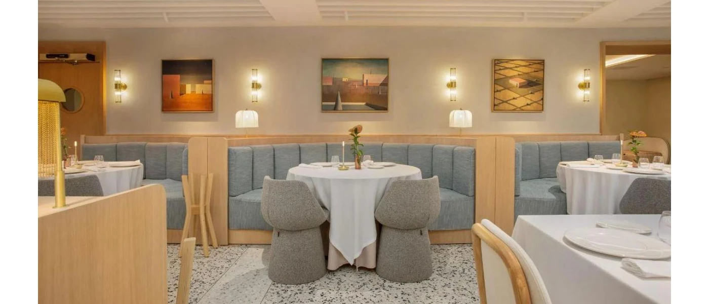 Modern restaurant with grey booth seating and chairs, a pebble-design floor, crisp white tablecloths and soft wall lighting with framed abstract pictures