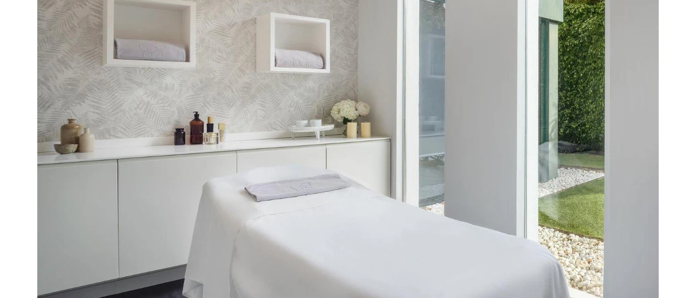 Treatment room with white bedding, vanity and walls, and full-length windows with gardens beyond