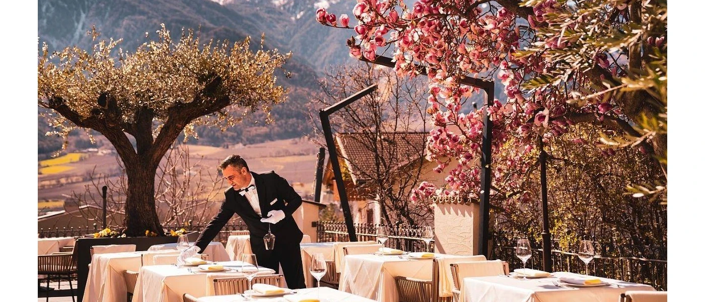 Waiter in black tie sets tables on an outdoor terrace dressed with cream tablecloths and surrounded by pink flowers, trees and mountains