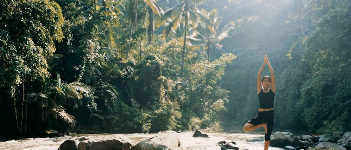 Woman in black active wear standing in a yoga pose in the jungle, with sun rays peering through the tree canopy