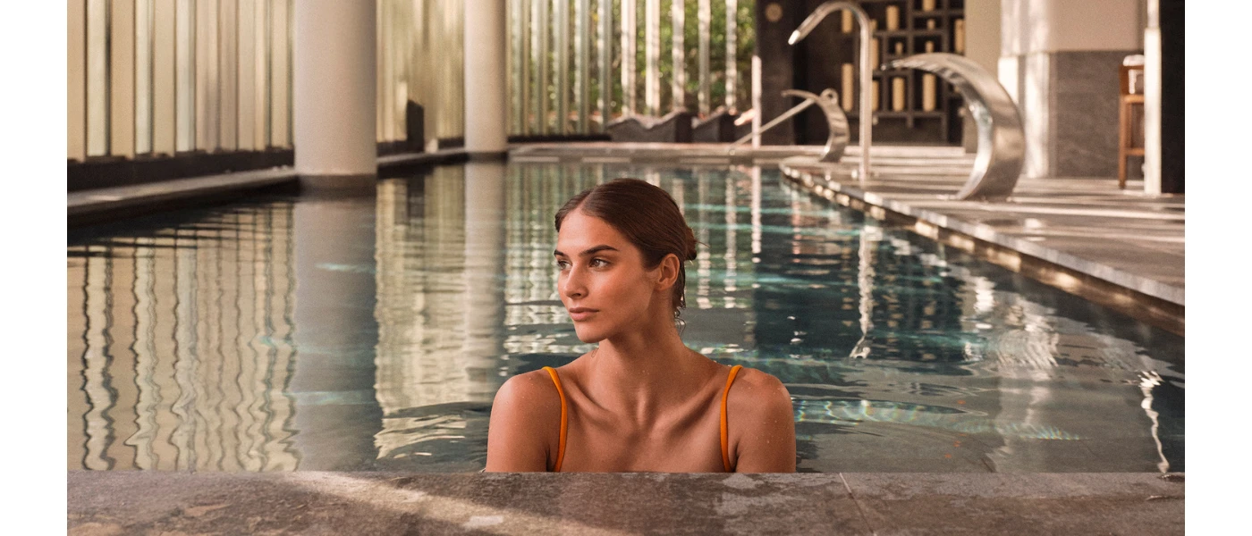 Woman in an orange swimsuit looks reflective in an indoor pool with silver water jets