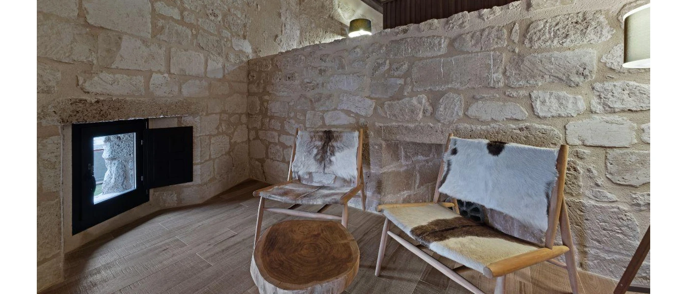Stone-walled room furnished with two wooden chairs made with animal fur, a small table made from a tree trunk, and a small black opening with a door serving as a window to outside