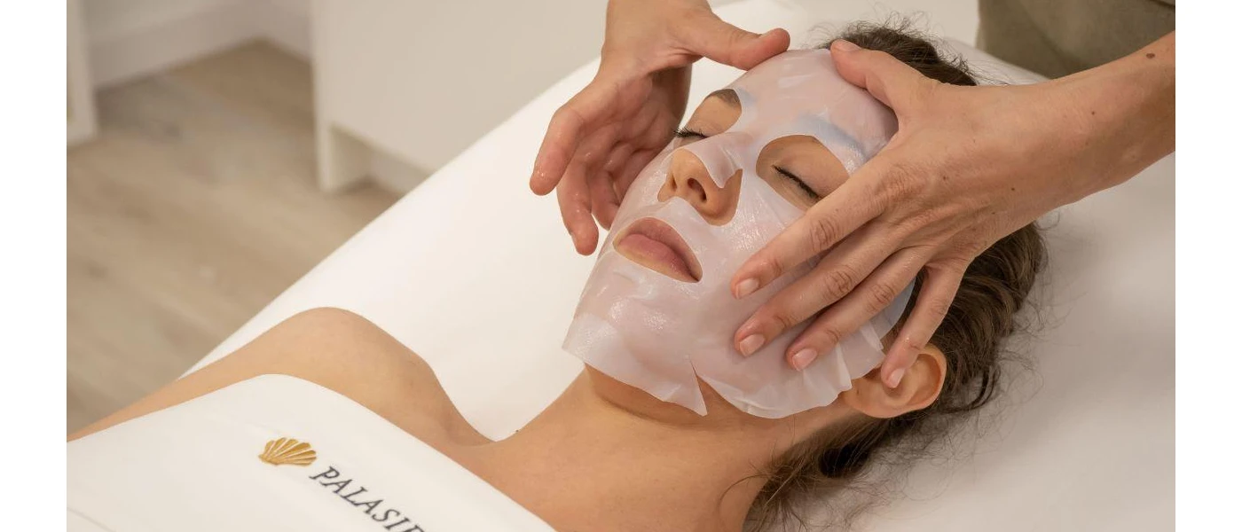 Woman lies with her eyes closed in a white towel as a therapist lies a sheet face mask on her