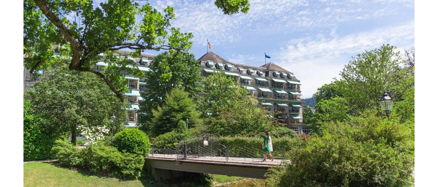 Woman walks over a bridge among gardens in front of a grand multi-storey building under a blue sky