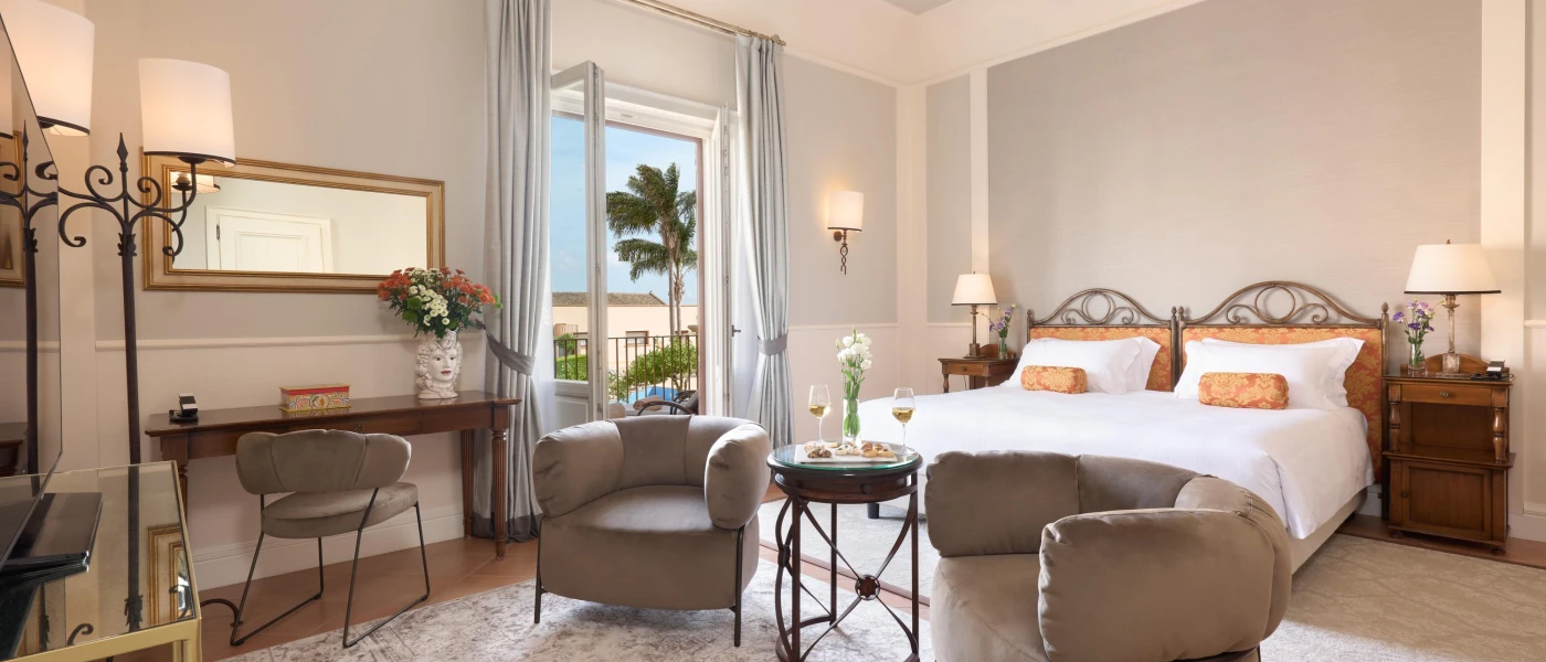 Elegant hotel room with grey walls and armchairs, white bedding with intricate headboards, dark-wood furniture and open balcony doors 