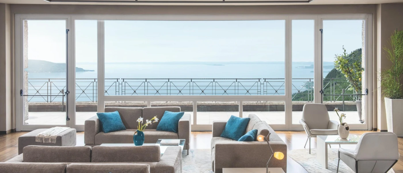 Neutral cushioned sofas with blue pillows and tables next to a huge window overlooking a terrace and Lake Garda