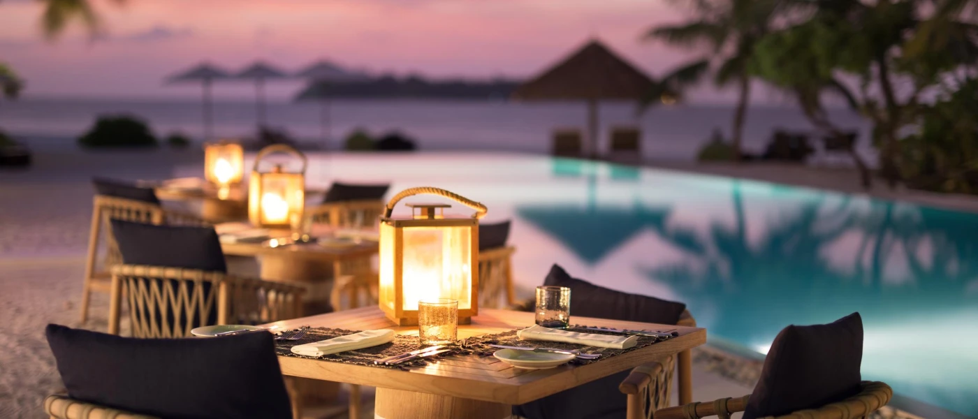 Wooden tables and chairs with black cushions set up for dinner by the poolside, topped with lanterns