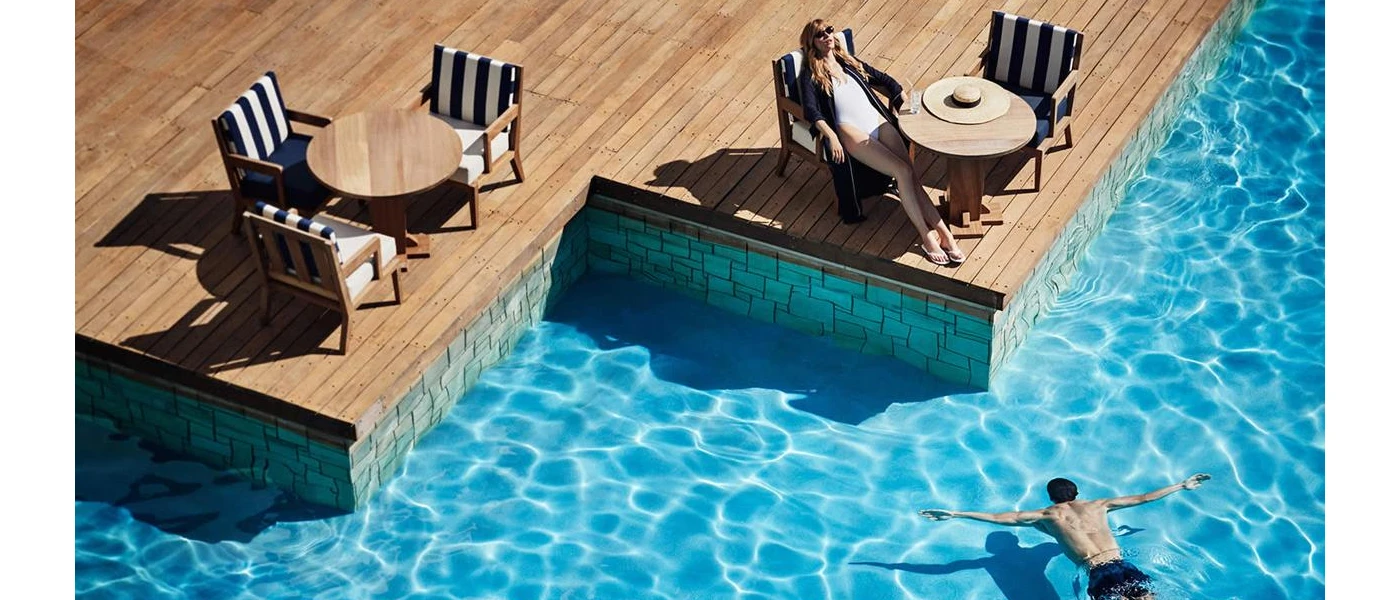 Woman in a white swimsuit and sunglasses relaxes on a blue and white-striped chair on a wooden terrace, as a man floats on his front in the neighbouring pool