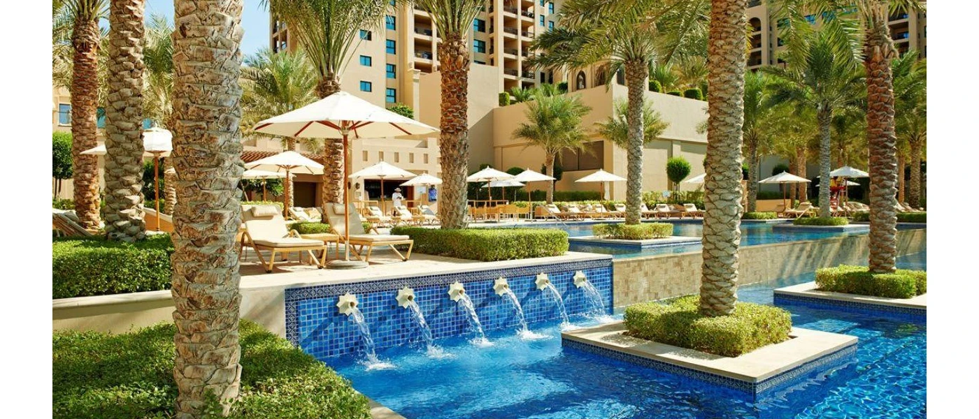 Multi-level pool with tiles and water jets in a terrace covered in palms, white parasols and loungers