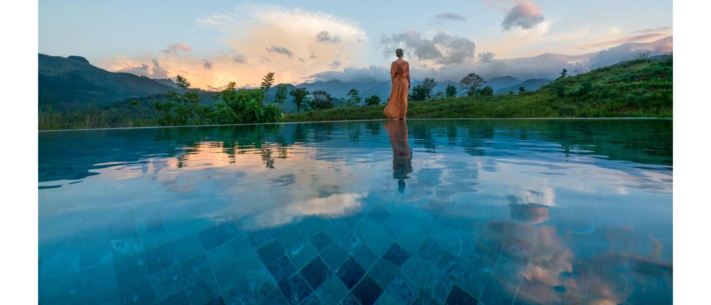 Woman standing in a full-length orange dress in front of a swimming pool overlooking mountains and rainforest as the sky darkens
