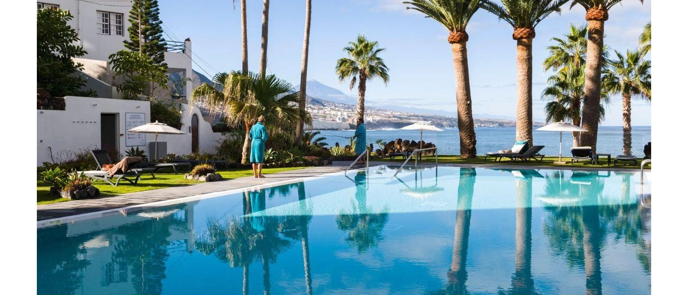 Guest in blue robe walking by a pool with palm trees and a sea view