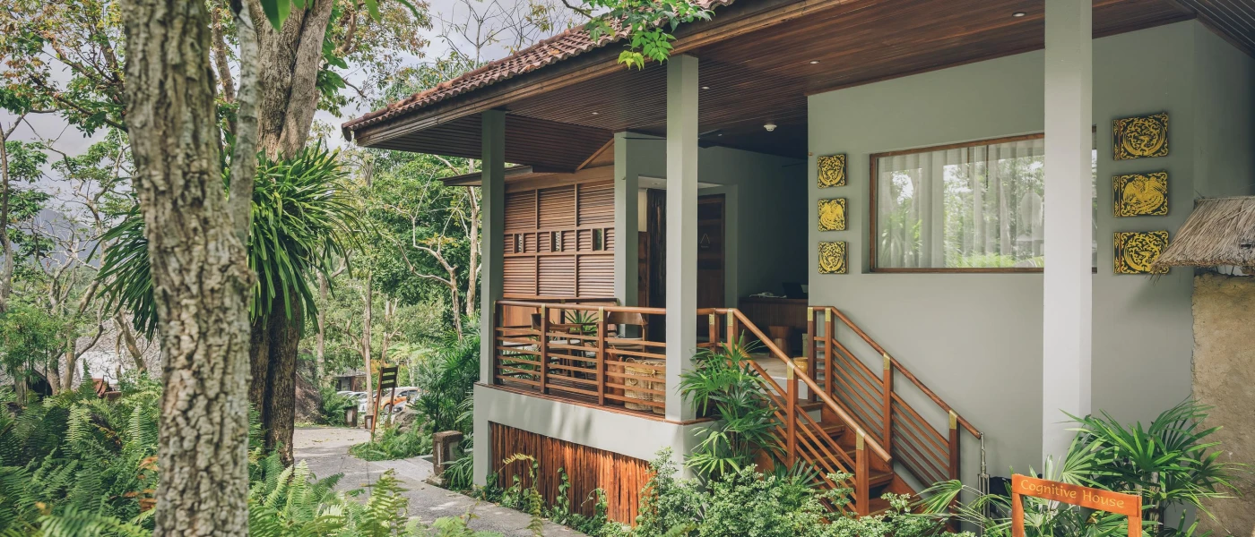 Low-rise villa with pillars, wood panelling, a terracotta tiled roof, gold icons on the wall and a surrounding of tropical greenery