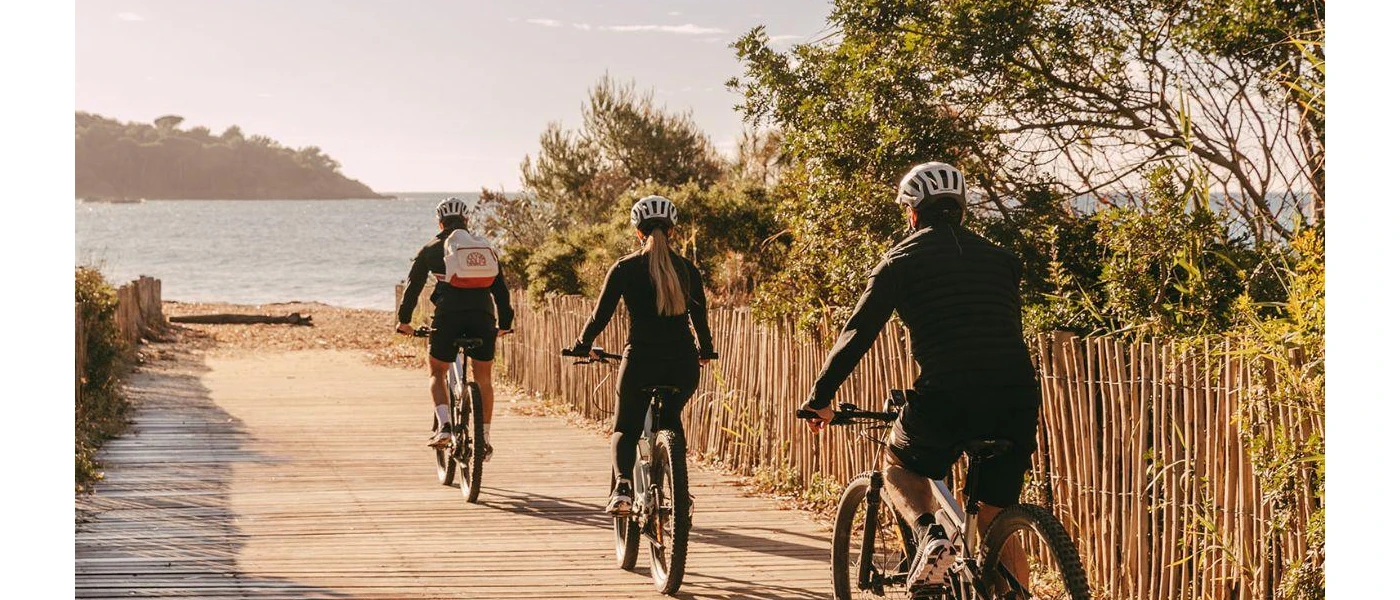 Three people in helmets and active wear cycle towards the sea along a pathway lined with greenery