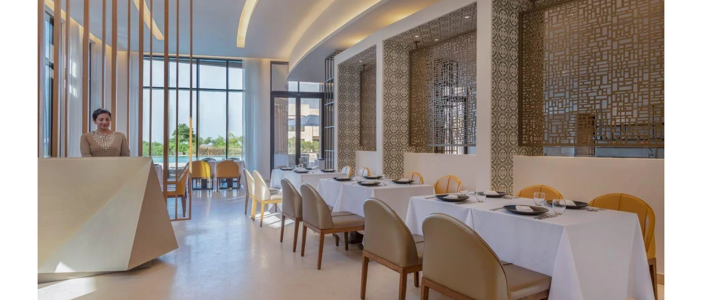 Restaurant with an intricate gold pattern on the walls, neutral leather seating, crisp white tablecloths and a smiling hostess standing behind a welcome desk