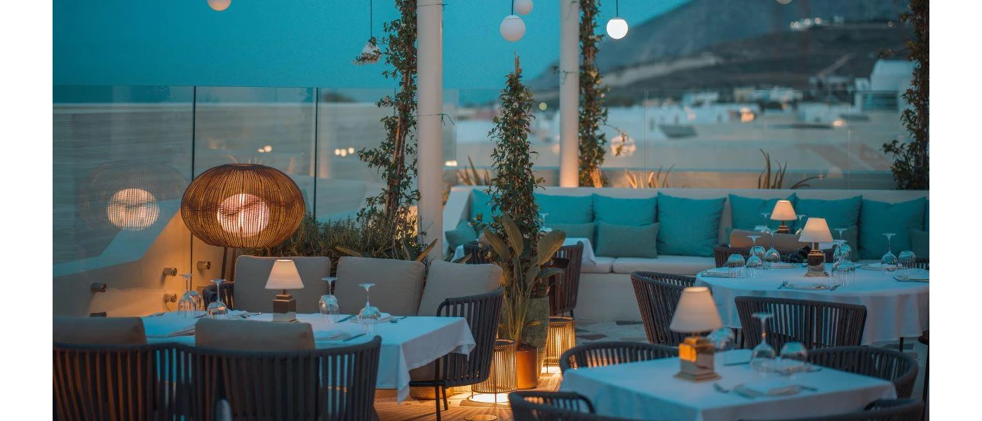 Stylish beach restaurant at night with glass wall surrounds, tables with white tablecloths, and sofas with teal cushions - lighting is soft and in rattan-style lights