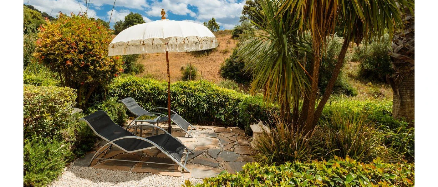 Stone patio with two black mesh loungers, a bohemian white umbrella with fringing, and tropical garden surroundings