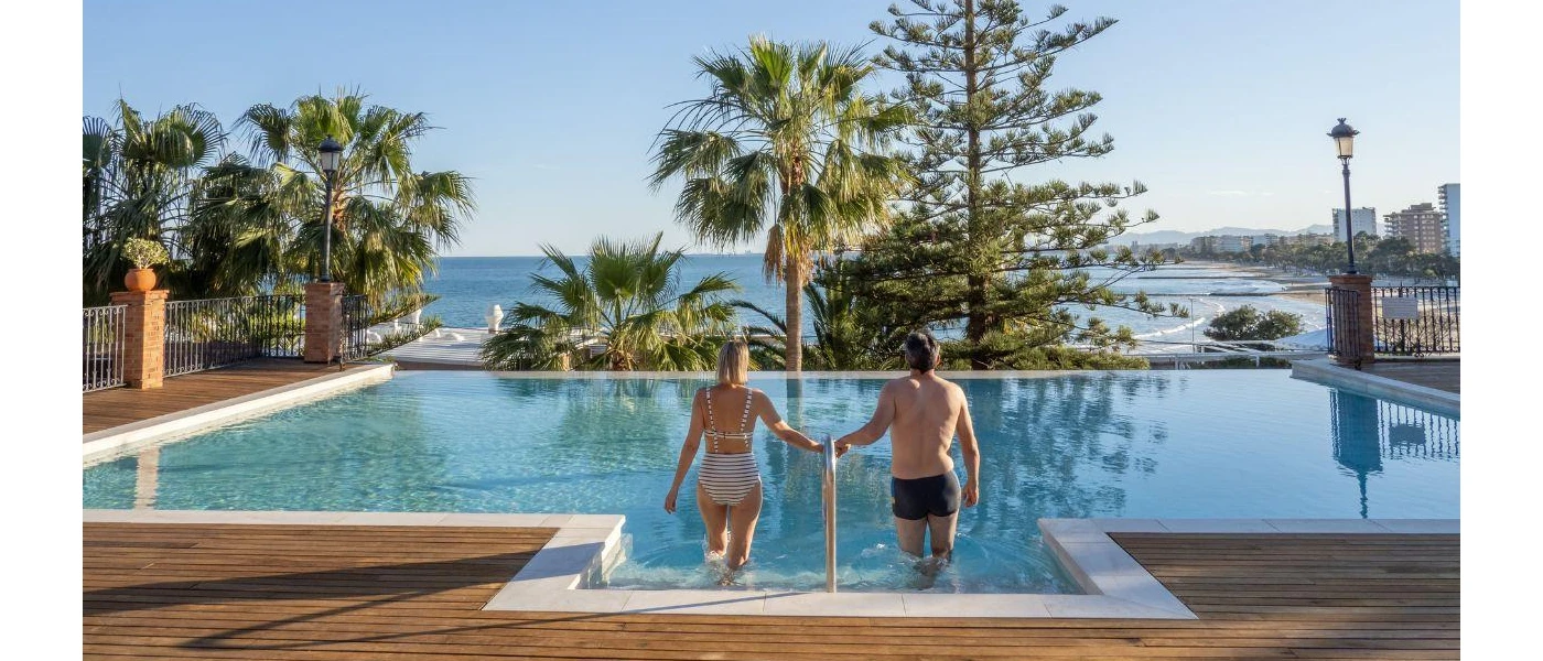 Woman in a striped black and white bikini and man in black trunks walk down steps into a pool set in a wooden terrace, with a view of greenery and the ocean beyond