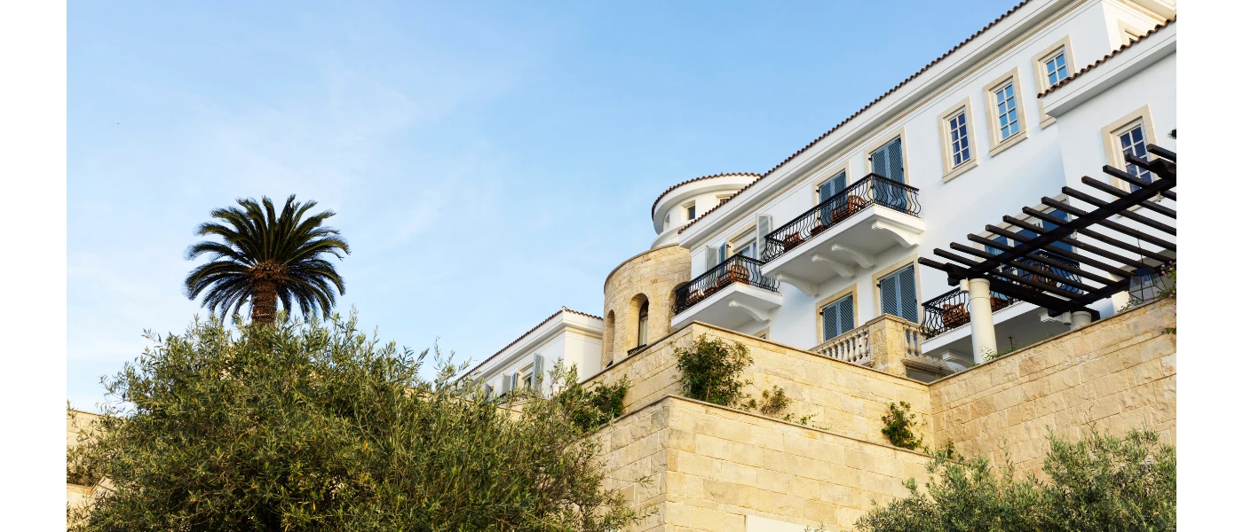 Building with white walls, black wrought-iron balconies and stone facades, fringed by a palm trees and wild flowers