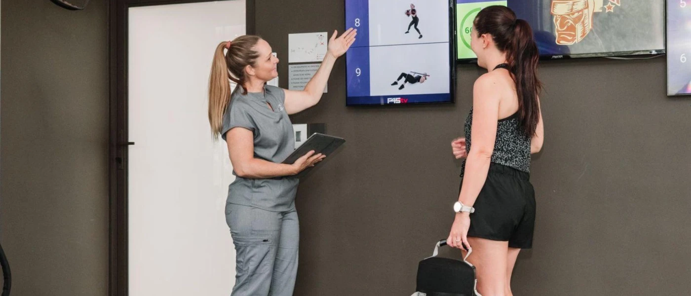 Female in grey uniform points at digital screens depicting workout animations as a woman in black active wear looks on